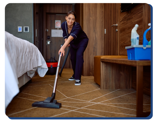 Hotel housekeeper vacuuming a carpeted guest room near the bed, with cleaning supplies in view, highlighting cleanliness and guest safety protocols.