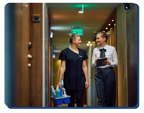 Two female hotel staff members walking down a hallway—one carrying cleaning supplies and the other holding a checklist—representing coordinated housekeeping and quality control.