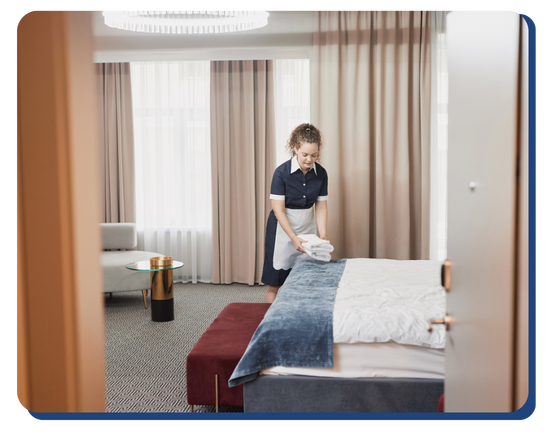Hotel housekeeper placing folded towels on a neatly made bed in a well-lit room, emphasizing daily cleaning and guest room upkeep.
