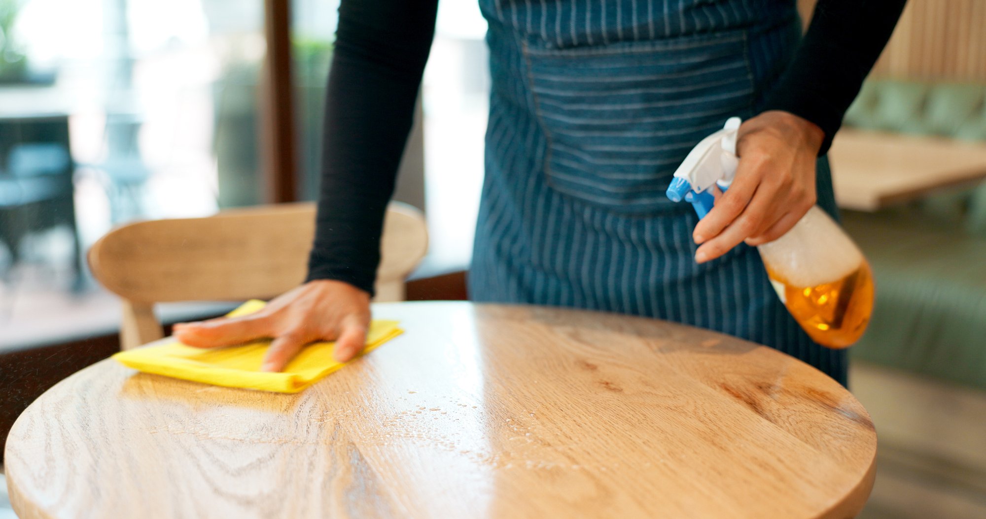 Staff member cleaning a wooden table with a yellow cloth and spray bottle as part of Toronto restaurant cleaning services. Staff member cleaning a wooden table with a yellow cloth and spray bottle as part of Toronto restaurant cleaning services.