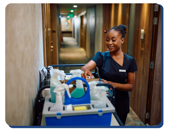 Smiling hotel housekeeper organizing cleaning supplies on a cart in the hallway, ready to begin room sanitation and housekeeping duties.