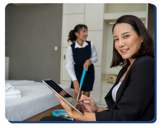 Hotel supervisor using a digital tablet to inspect a room while a housekeeper cleans in the background, highlighting quality assurance in hotel cleaning services.