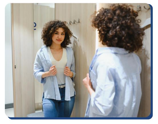 Woman trying on clothes in a clean retail fitting room, standing in front of a mirror with wooden walls and hooks, emphasizing cleanliness and customer comfort.