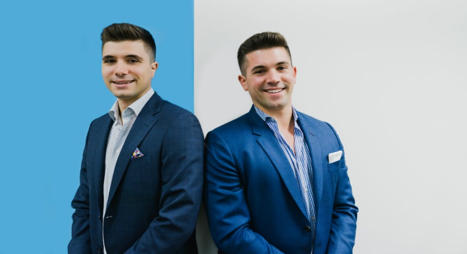 Two young men in blue suits stand side by side, smiling, against a background split vertically between light blue and white.