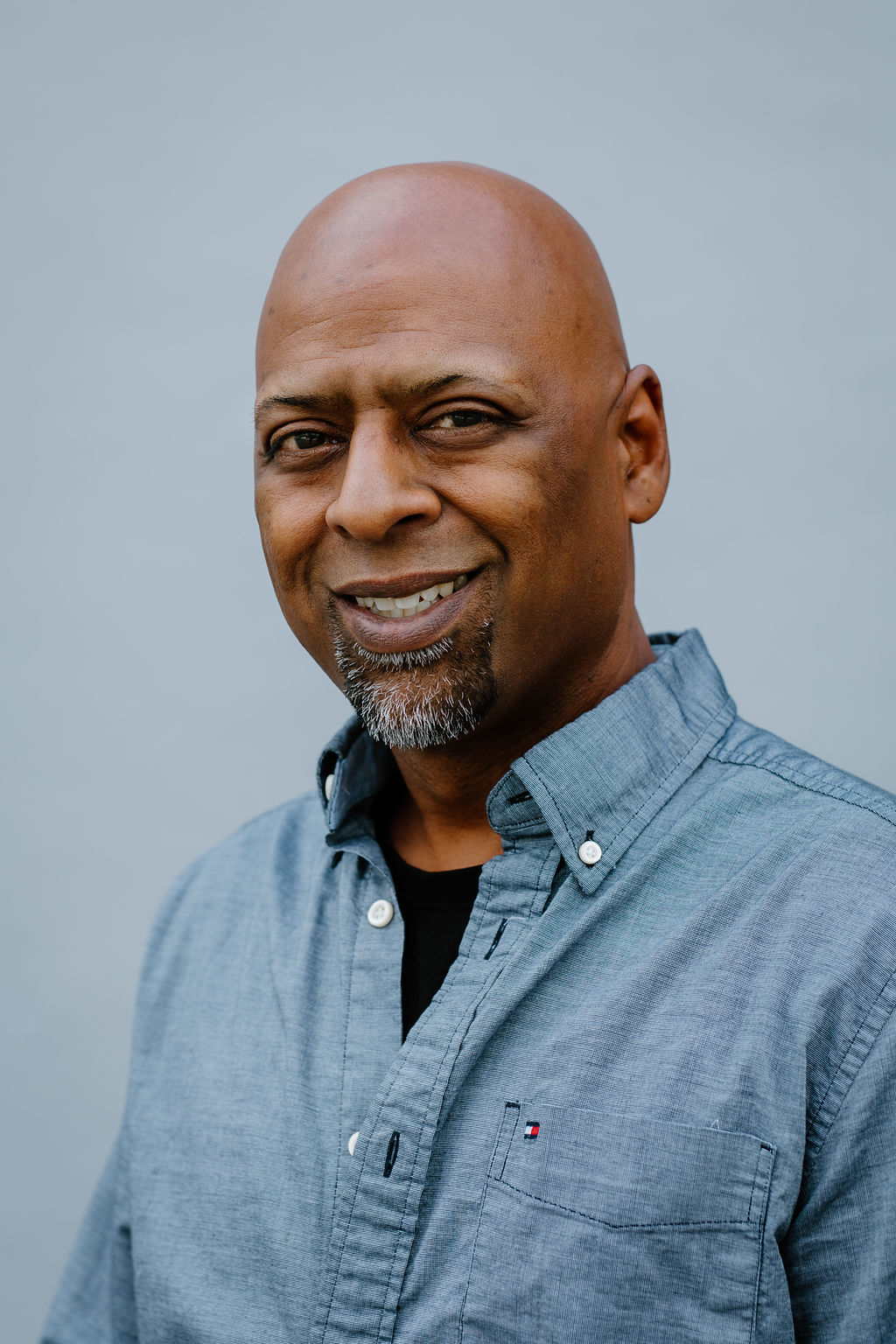 A bald man with a goatee smiles at the camera while wearing a blue button-up shirt over a black t-shirt, standing in front of a plain light gray background.