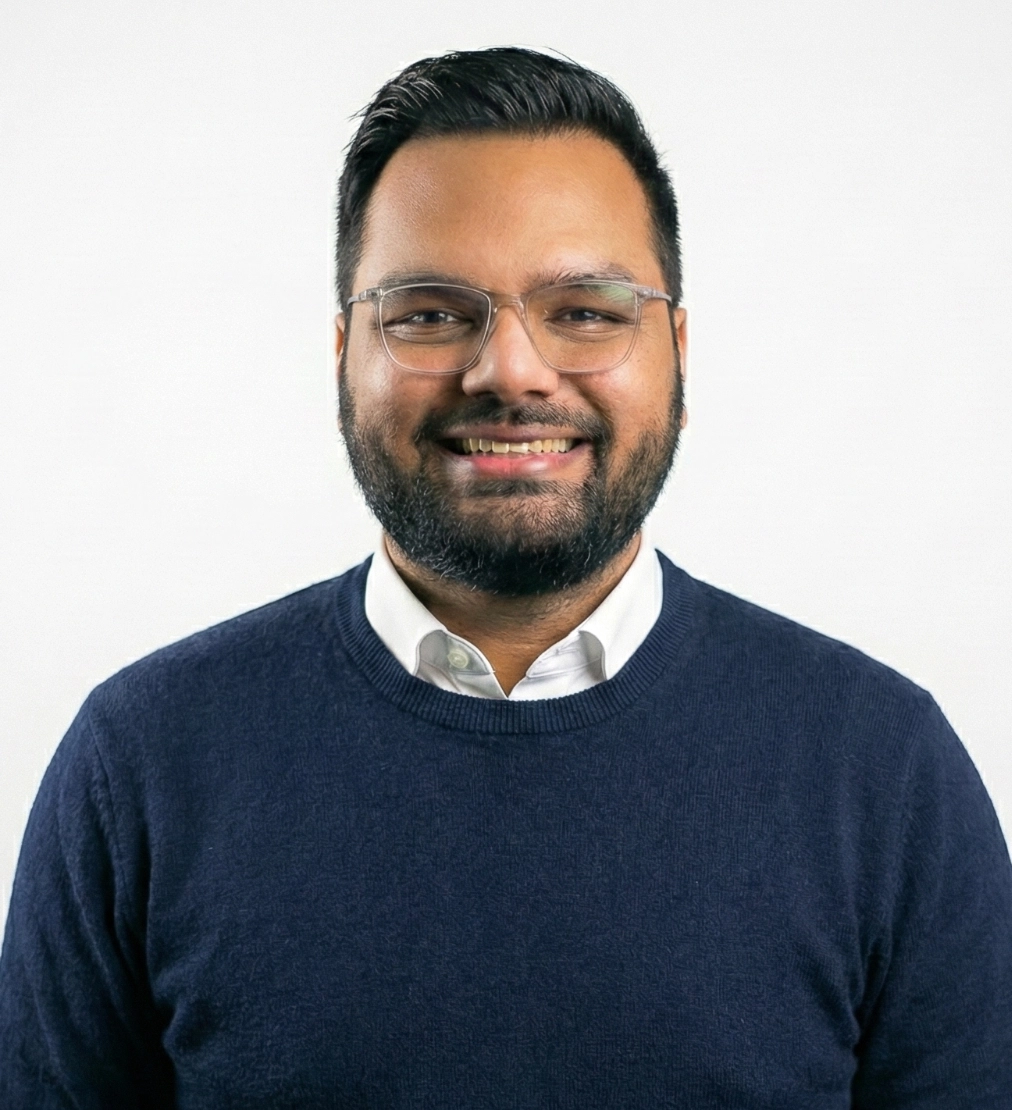 A man with short dark hair, glasses, and a beard smiles at the camera. He is wearing a navy blue sweater over a white collared shirt, standing against a plain white background.