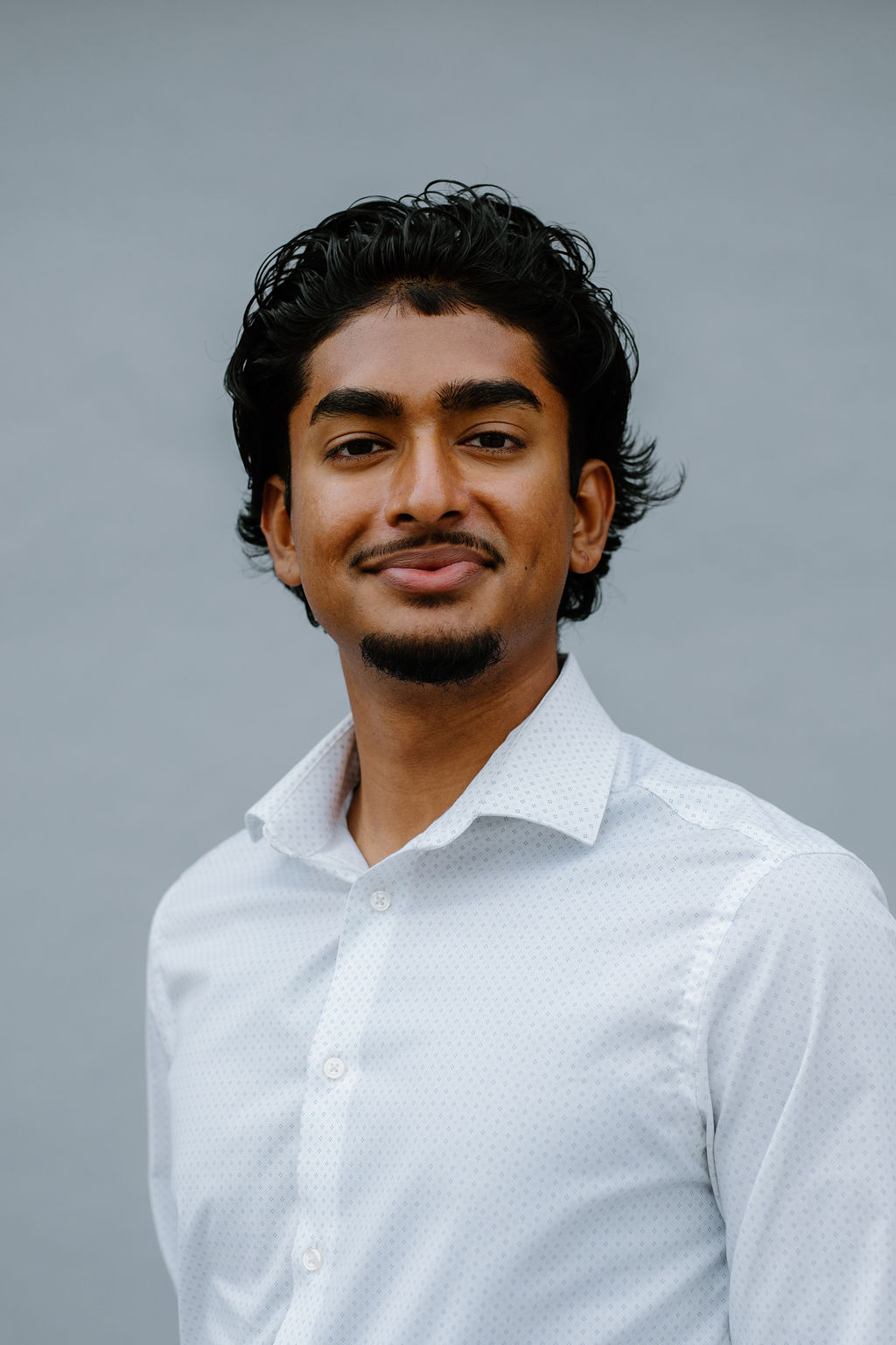 A young man with dark curly hair, a trimmed beard, and mustache, wearing a white button-up shirt, stands in front of a plain gray background, smiling slightly at the camera.