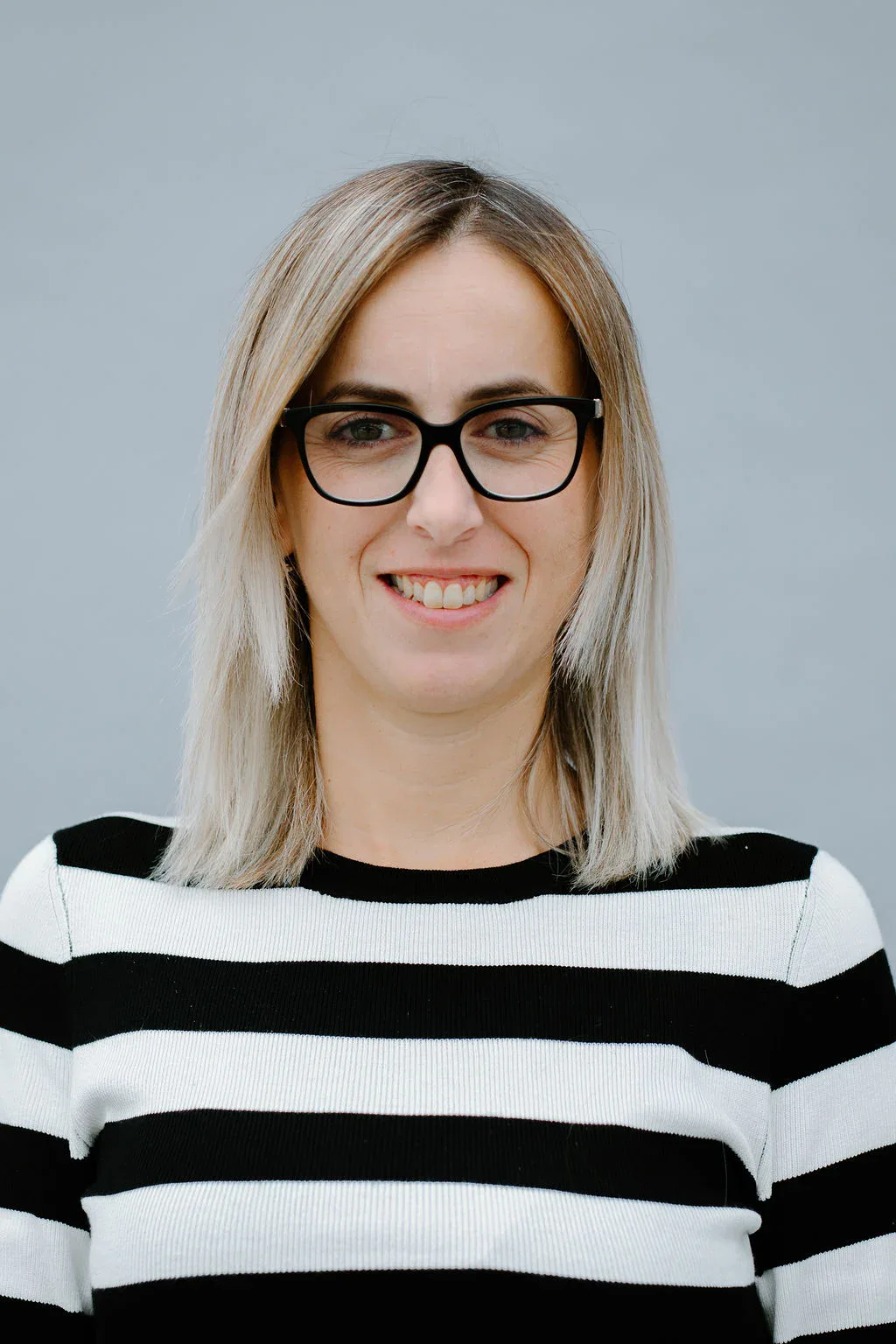 A woman with straight, shoulder-length blonde hair, wearing black-rimmed glasses and a black-and-white striped top, smiles at the camera against a plain light gray background.