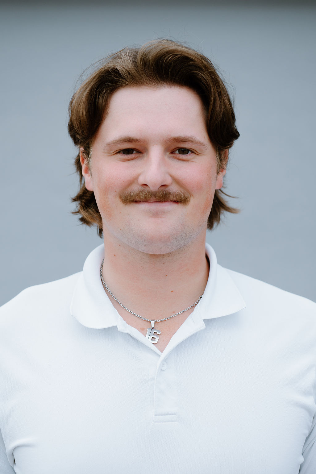 A man with medium-length brown hair and a mustache, wearing a white collared shirt and a silver necklace, stands in front of a plain gray background, smiling slightly.
