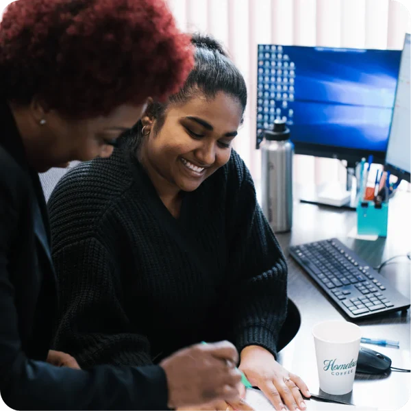 Two women smiling and working together at a desk with a computer, documents, and a coffee cup, in a bright office setting.
