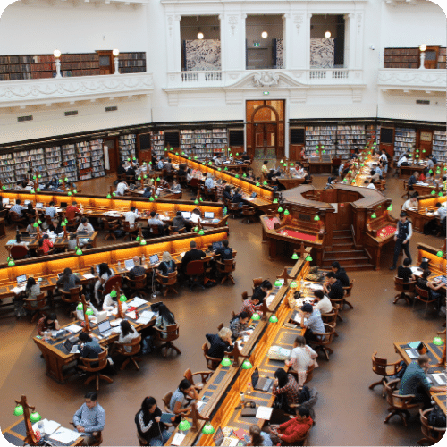 A spacious library reading room with people studying at large wooden tables, surrounded by bookshelves and illuminated by green desk lamps. The room has high ceilings and large arched windows.