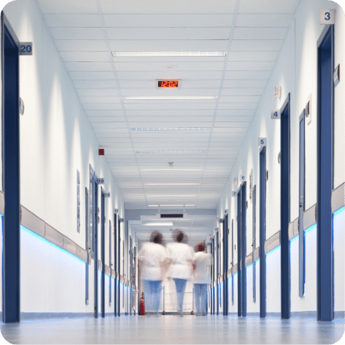 A hospital corridor with white walls and blue accents, lined with numbered doors on both sides. Three people in white uniforms walk away from the camera toward the end of the hallway.