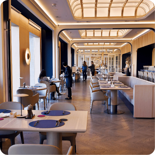 A modern, elegant restaurant with high ceilings, stylish lighting, and spacious seating. A few patrons are dining, while staff serve and set tables. Large windows let in natural light, creating a warm, inviting atmosphere.