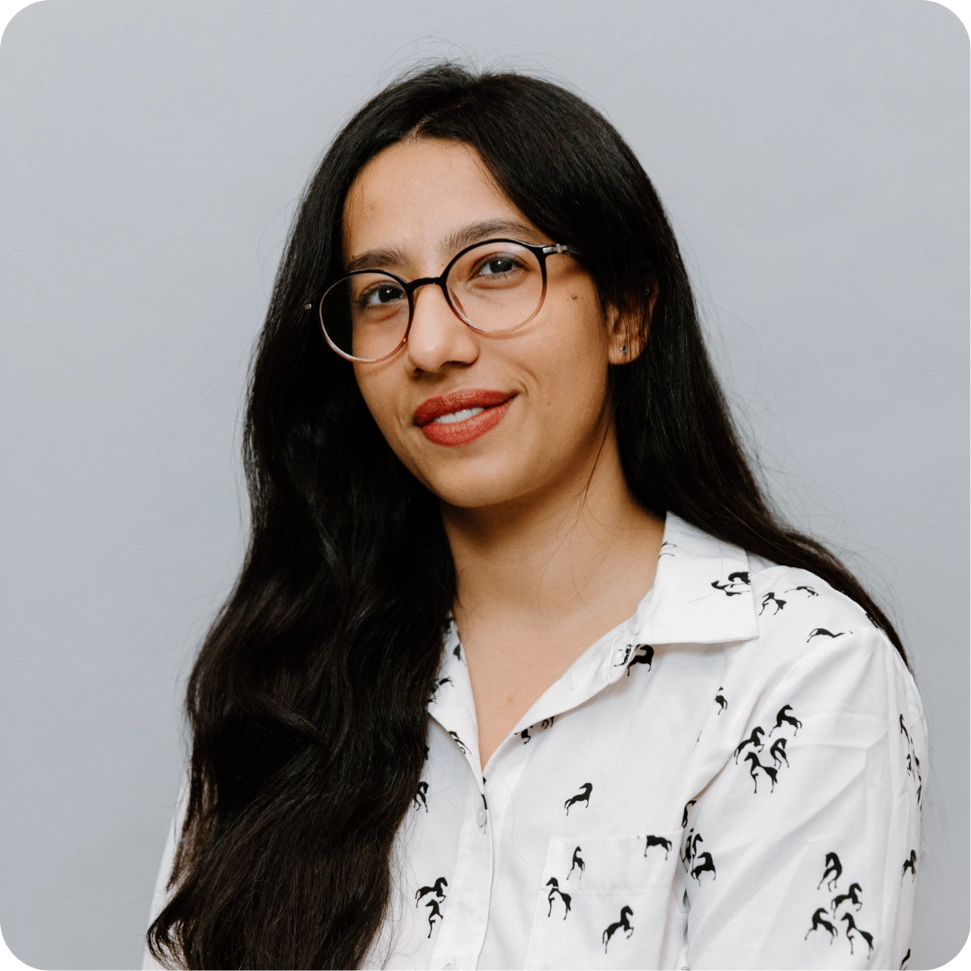 A woman with long dark hair and glasses smiles slightly, wearing a white shirt with black horse patterns, posing against a plain light gray background.