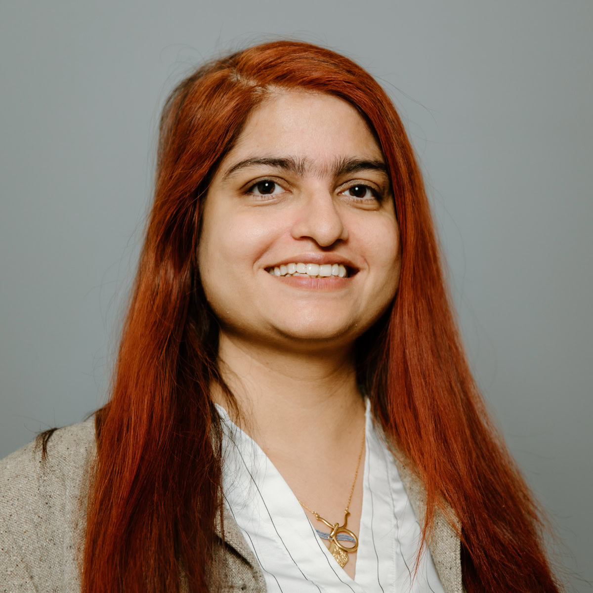 A woman with long, straight reddish-brown hair smiles at the camera. She is wearing a beige jacket over a white blouse and a gold necklace, standing in front of a plain grey background.