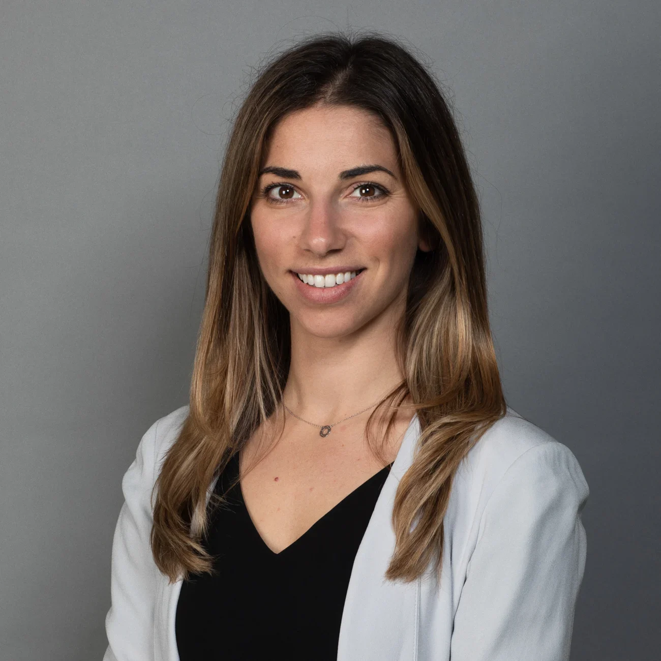 A woman with long, straight brown hair wearing a light gray blazer over a black top, smiling at the camera against a plain gray background.
