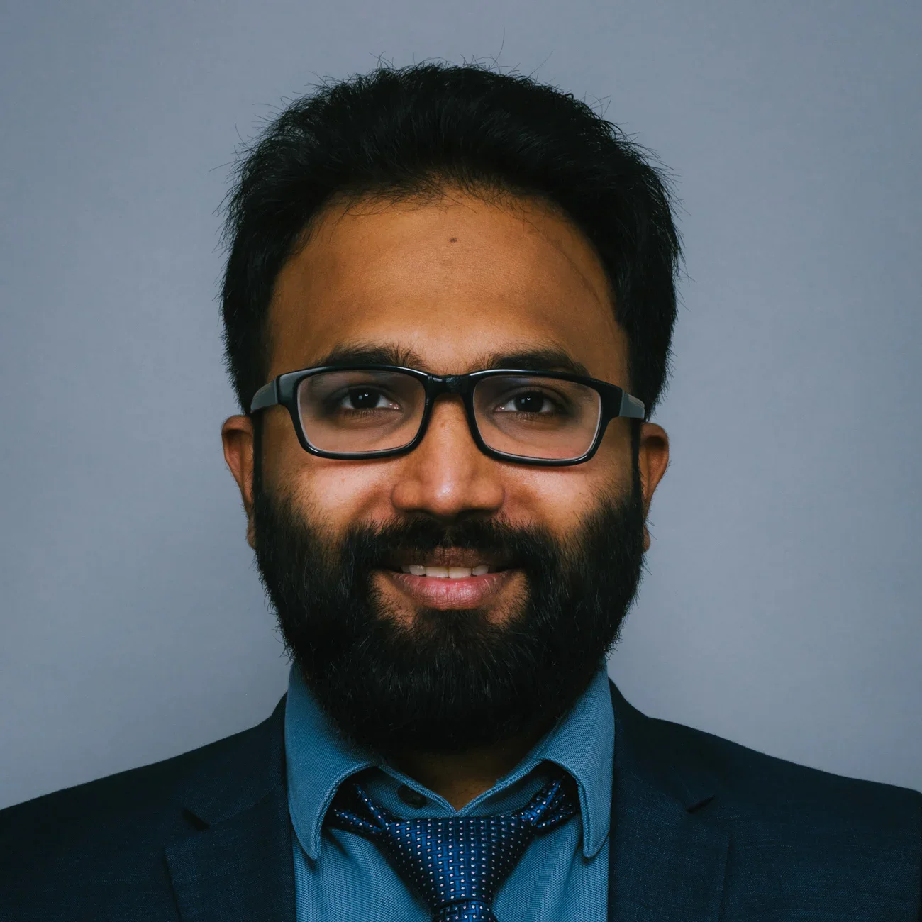 A man with short black hair, a full beard, and glasses is smiling. He is wearing a dark suit jacket, a blue shirt, and a blue patterned tie. The background is plain and gray.