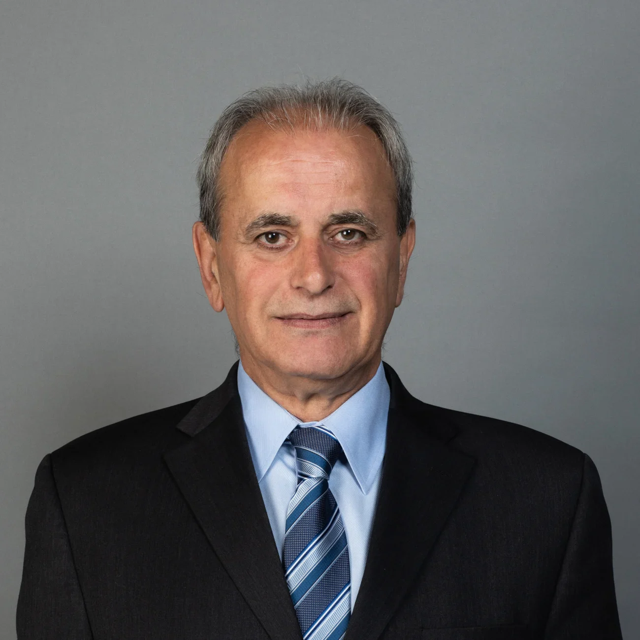 An older man with gray hair wearing a black suit, light blue shirt, and striped blue tie, smiling slightly, posed in front of a plain gray background.