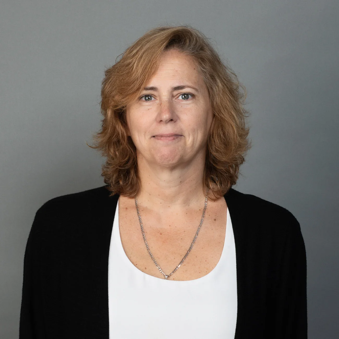 A woman with wavy, shoulder-length light brown hair wears a white top, a black cardigan, and a silver necklace, standing in front of a plain gray background, smiling softly at the camera.