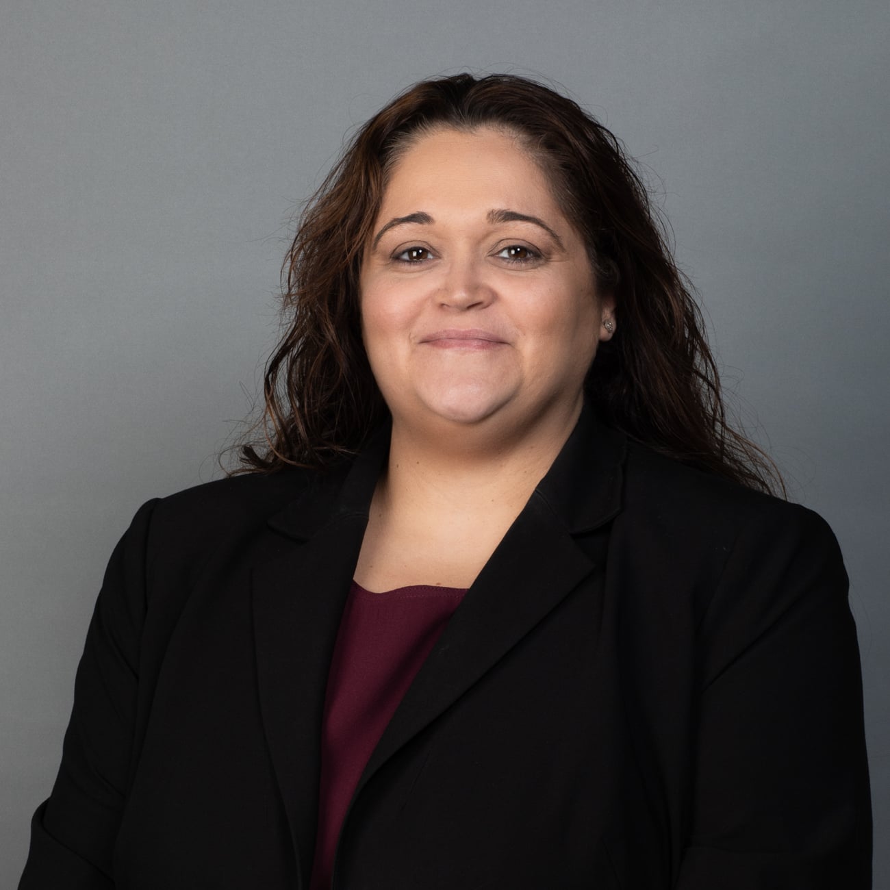 A woman with long brown hair, wearing a black blazer over a maroon top, smiles softly at the camera against a plain gray background.