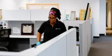 A woman wearing a colorful headband and gloves cleans an office cubicle. She is focused on her work, and the office has gray cubicle walls, framed certificates, and artwork in the background.