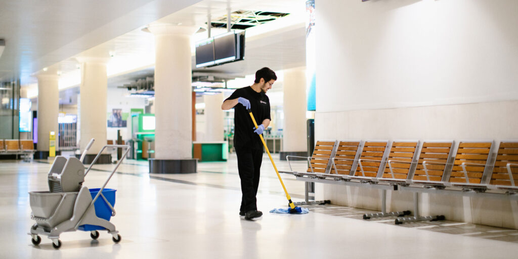 A person wearing gloves is mopping the floor in a spacious, bright indoor area with benches and a cleaning cart nearby. The space appears to be a public waiting area, possibly an airport or train station.