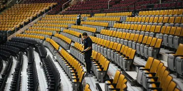 A man stands alone among rows of empty yellow and brown stadium seats, looking down at something in his hands. The arena is deserted, with only one other person visible in the distance.