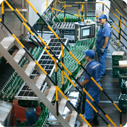 Workers in blue uniforms operate machinery on a factory floor with green bottles on a conveyor belt, surrounded by metal railings and industrial equipment.