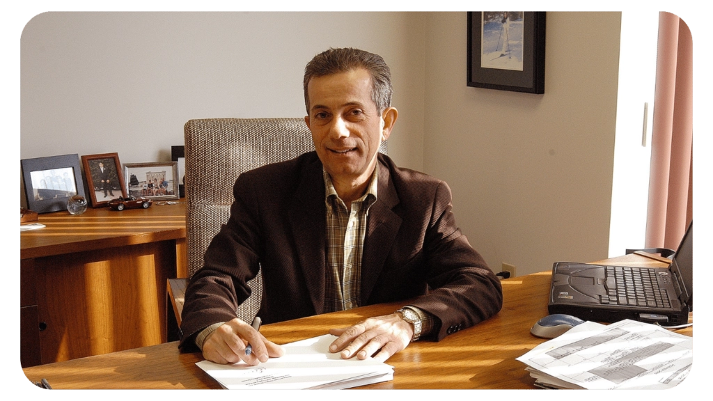 A man in a brown jacket sits at a wooden desk with papers and a laptop, smiling at the camera. The office has framed photos, a chair, and a window letting in natural light.