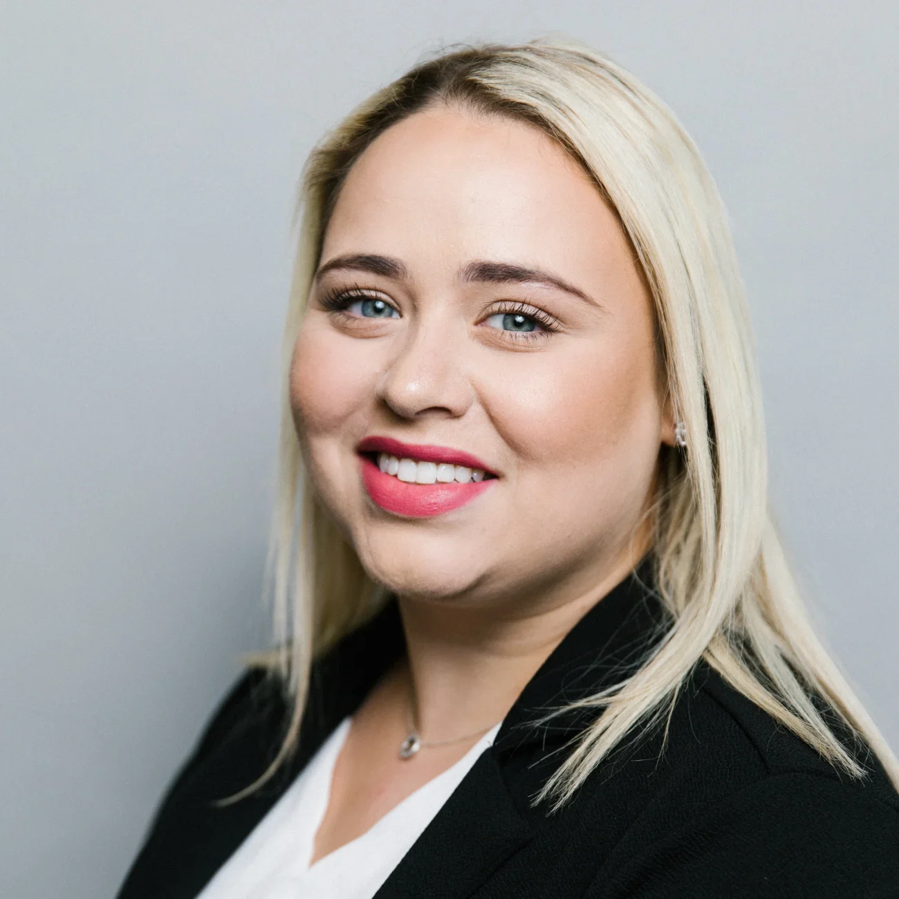 A woman with straight blonde hair smiles at the camera. She is wearing a black blazer over a white top, has light skin, pink lipstick, and is posed against a plain light gray background.