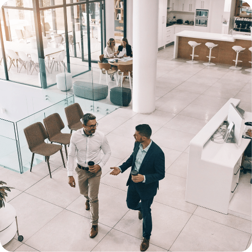 Two men in business attire walk and talk in a modern, open office space holding coffee cups. In the background, three women sit at a table, and the kitchen area is visible with bar stools and cabinets.