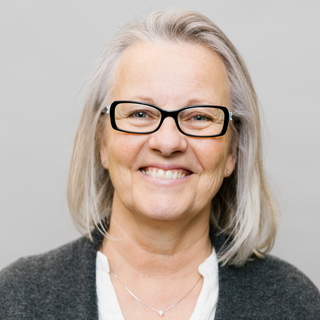 Smiling older woman with straight, shoulder-length gray hair, wearing black-framed glasses, a white blouse, and a dark gray cardigan, standing in front of a plain light gray background.
