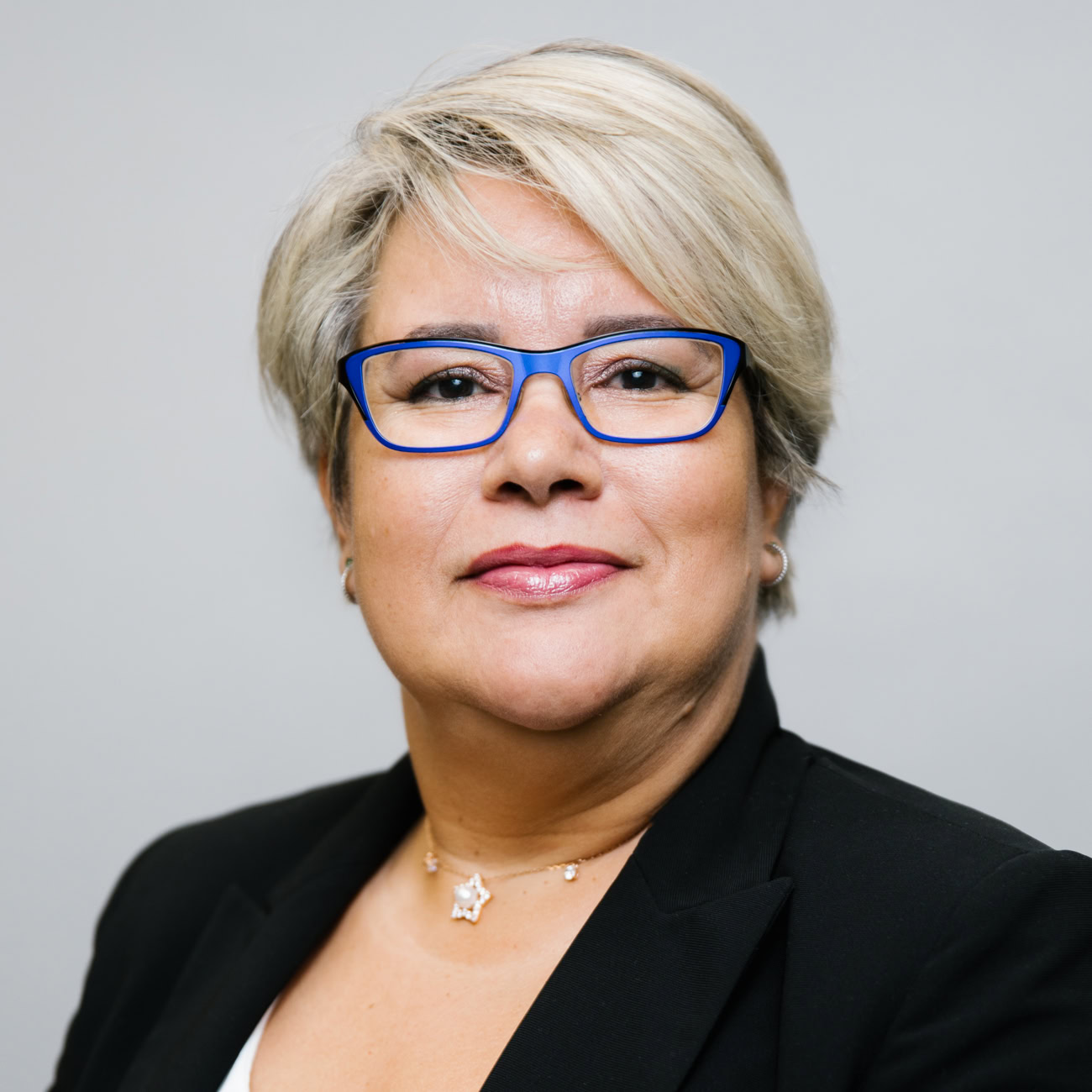 A woman with short blonde hair, wearing blue-framed glasses, a black blazer, a pearl earring, and a star necklace, poses against a plain light gray background.