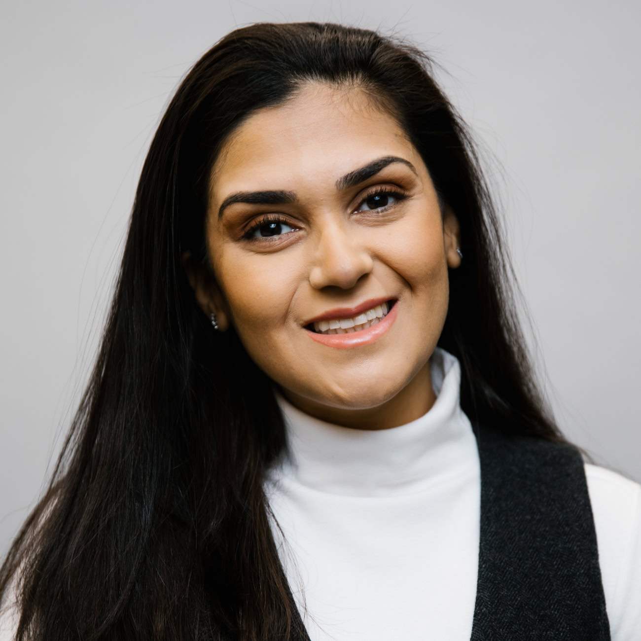 A woman with long dark hair, wearing a white turtleneck and dark vest, smiles in front of a plain light gray background.