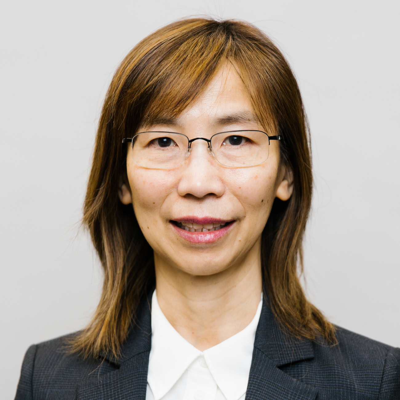 A woman with straight, shoulder-length brown hair and glasses, wearing a dark blazer and a white collared shirt, is smiling slightly against a plain light gray background.