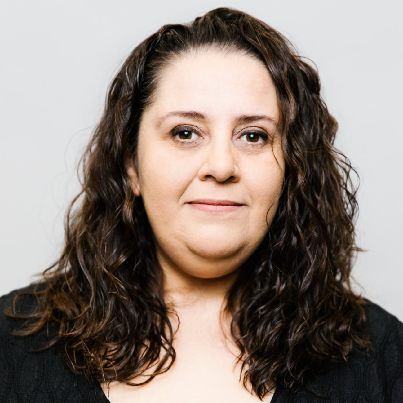 A woman with long, wavy brown hair and a light complexion faces the camera, smiling softly. She is wearing a black top and is posed against a plain, light gray background.