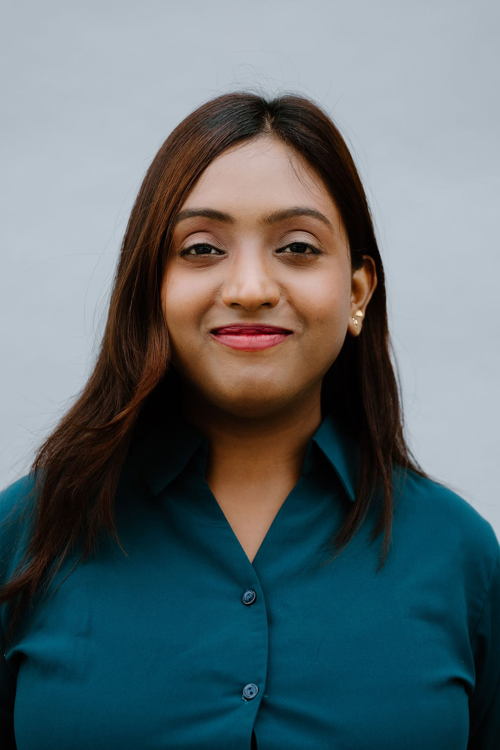 A woman with long brown hair, wearing a teal button-up shirt, stands in front of a plain light gray background, smiling gently at the camera.