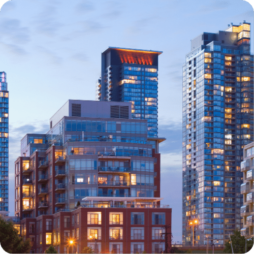 Modern high-rise apartment buildings with glass windows and balconies, illuminated from within at dusk under a partly cloudy sky.