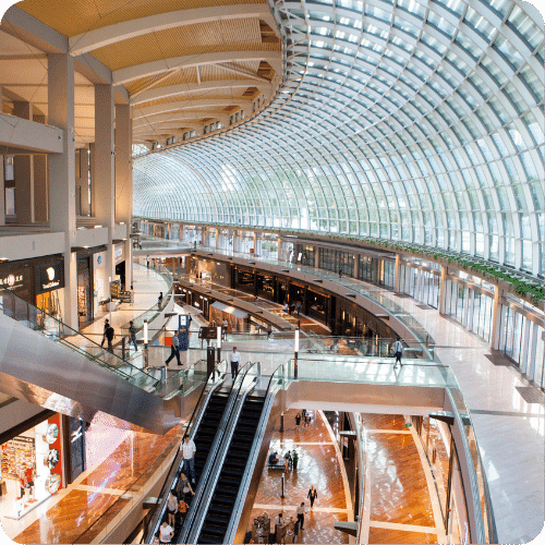 A spacious, modern shopping mall with glass arched ceilings, multiple floors, escalators, and people walking along polished floors and shopfronts. Natural light floods the interior through large windows.