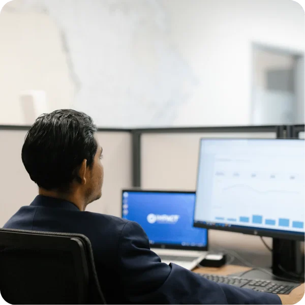 A man in a suit sits at a desk in an office, looking at charts and graphs on a large computer monitor, with a laptop and divider panels in the background.
