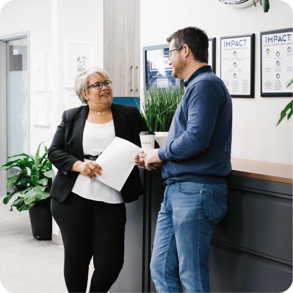 A woman in a black blazer holds papers and smiles while talking to a man in a blue sweater holding a coffee cup. They stand in a modern office with plants and posters on the wall.