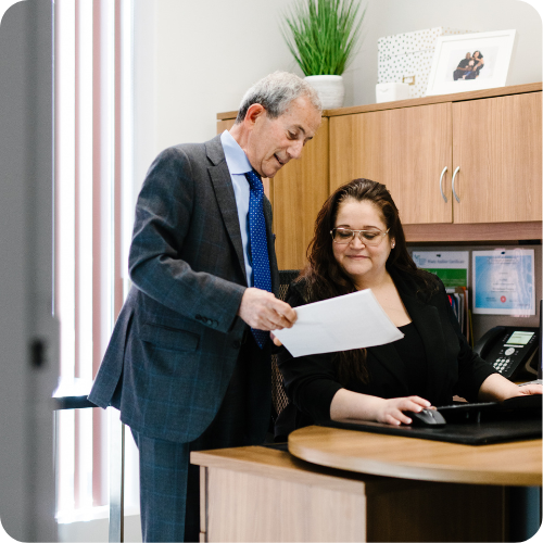 A man in a suit shows a document to a woman seated at an office desk. The woman looks at the paper while using a computer. Office decor and family photos are visible in the background.