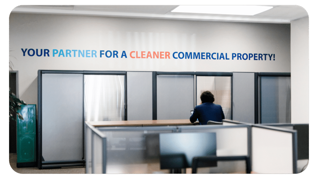 A person in a suit sits alone at a desk in an office. On the wall behind them is a sign that reads, YOUR PARTNER FOR A CLEANER COMMERCIAL PROPERTY! in bold, colorful text.