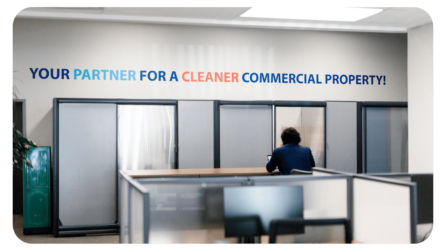 A person in a suit sits alone at a desk in an office. On the wall behind them is a sign that reads, YOUR PARTNER FOR A CLEANER COMMERCIAL PROPERTY! in bold, colorful text.