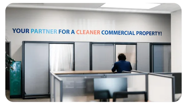 A person sits alone at a desk in a modern office space. The wall behind them displays the slogan, YOUR PARTNER FOR A CLEANER COMMERCIAL PROPERTY! in bold, colorful letters.