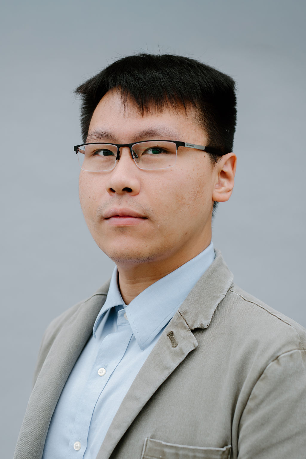 A man with short black hair and glasses wearing a light blue collared shirt and a light gray blazer stands against a plain gray background, looking slightly to the side.