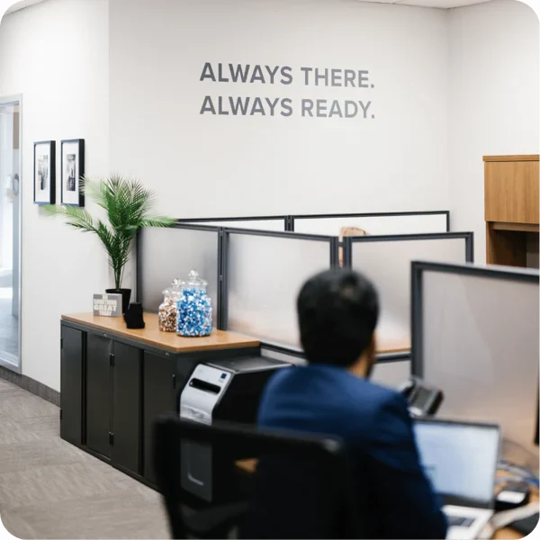 A modern office with cubicles, a person working on a laptop, a potted plant, and jars of candy. The wall displays the words ALWAYS THERE. ALWAYS READY. in large letters.