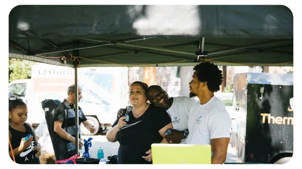 Four people stand and interact under a tent at an outdoor event. One woman holds a microphone, two men in white shirts stand beside her smiling, and another woman stands on the left using her phone. A van and trailer are visible in the background.