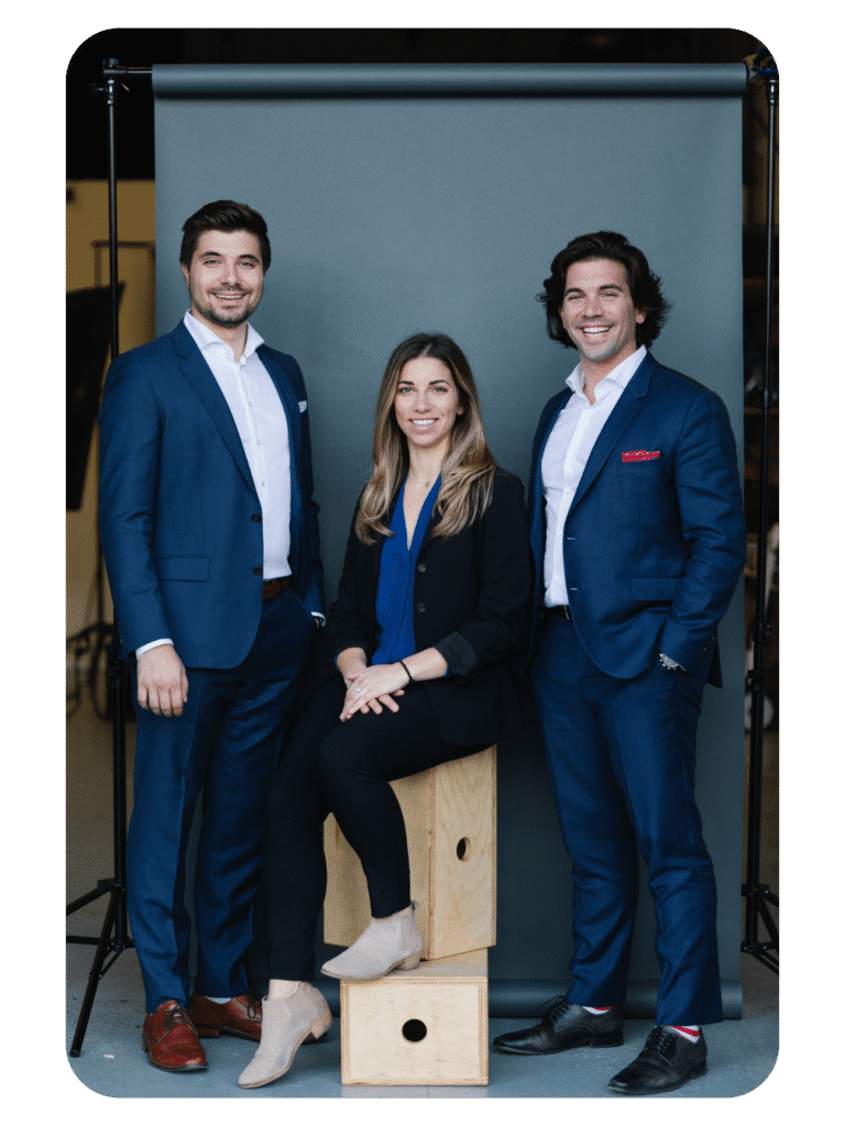 Three people in business attire pose together against a gray backdrop; two men in blue suits stand on either side of a woman seated on a wooden box, all smiling at the camera.