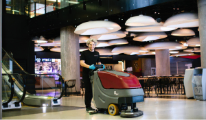 A person in black clothing and blue gloves operates a large red and gray floor cleaning machine in a modern, spacious indoor area with escalators, tables, and unique ceiling lights.
